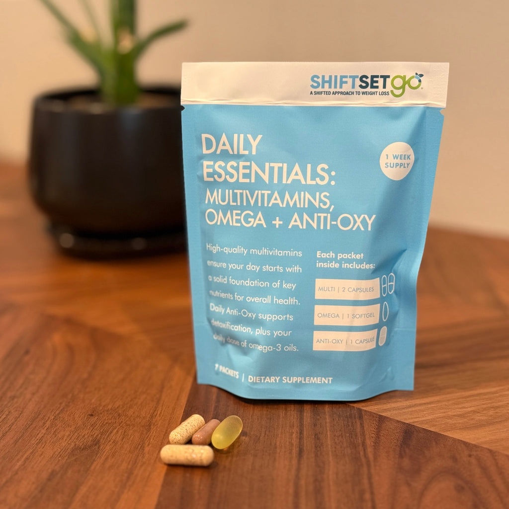 A weekly pouch labeled 'Daily Essentials: Multivitamins Omega + Anti-Oxy' from ShiftSetGo, containing packets of supplements, displayed on a wooden surface with a plant in the background.