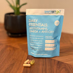 A weekly pouch labeled 'Daily Essentials: Multivitamins Omega + Anti-Oxy' from ShiftSetGo, containing packets of supplements, displayed on a wooden surface with a plant in the background.