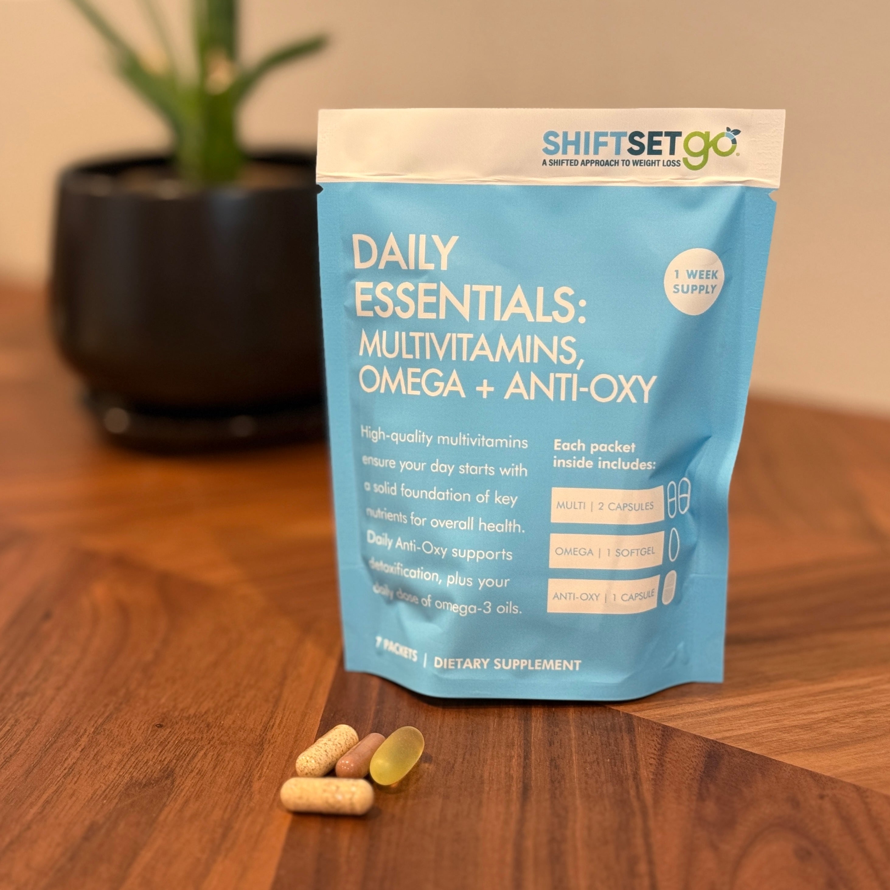 A weekly pouch labeled 'Daily Essentials: Multivitamins Omega + Anti-Oxy' from ShiftSetGo, containing packets of supplements, displayed on a wooden surface with a plant in the background.
